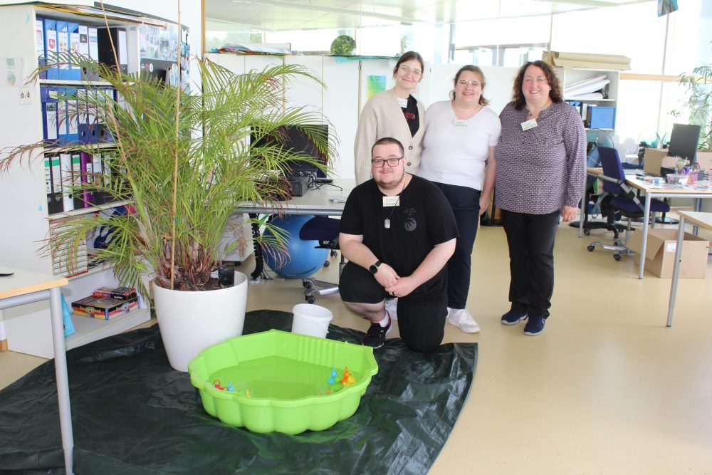 Vier Auszubildende in ihrem Ausbildungsbüro; außerdem sind eine Palme und ein Planschbecken mit Enten zu sehen