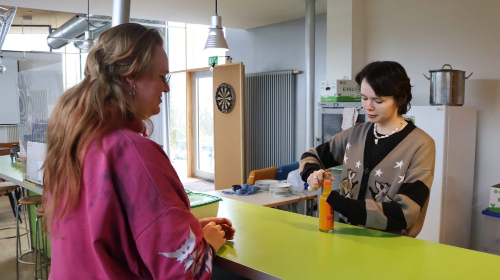 eine Personen steht vor, eine hinter einer Bistrotheke; die Person hinter der Theke öffnet eine Flasche Limonade