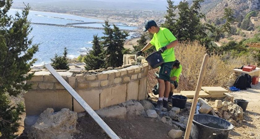 Ein junger Mann arbeitet an einer Natursteinmauer; im Hintergrund sieht man das Meer