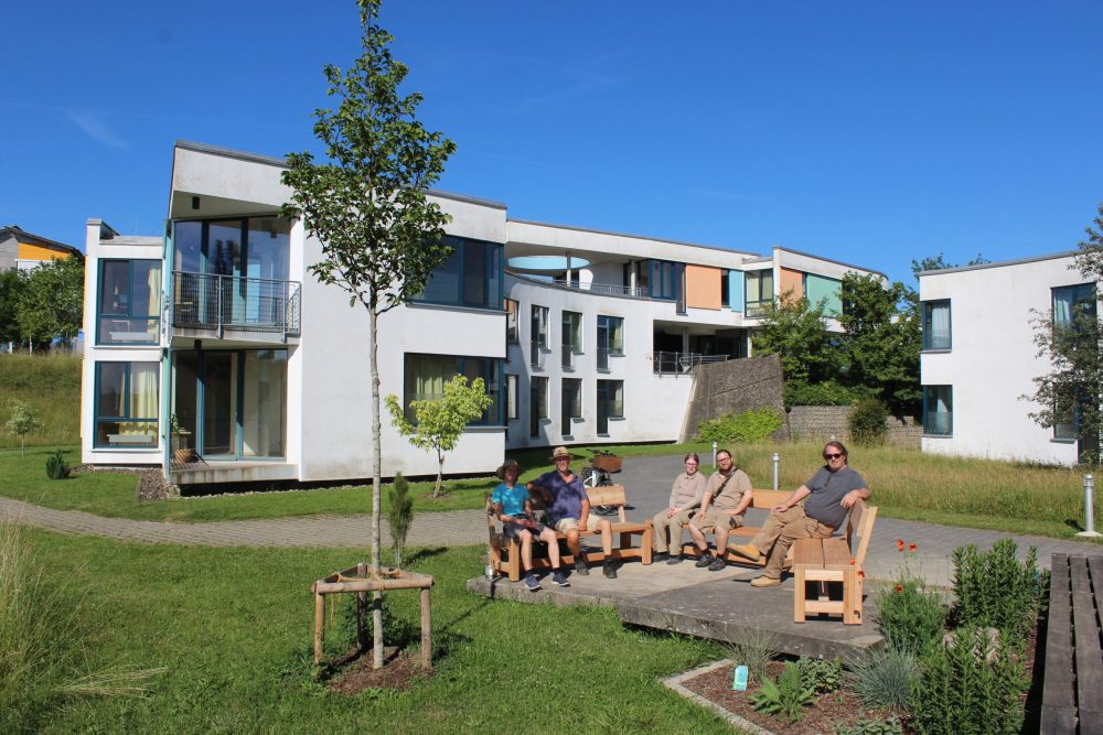 die Kolleg*innen vom Bereich GaLa-Bau, die beiden Azubis aus der Schreinerei und ihr Ausbilder auf den Sitzbänken an der Gedenkstätte