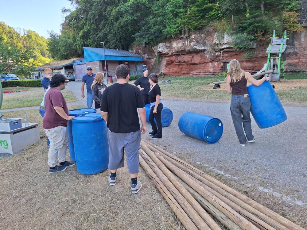 Eine Gruppe Teilnehmende bauen ein Floss aus blauen Tonnen, Holzstämmen, Seilen und Spanngurten.