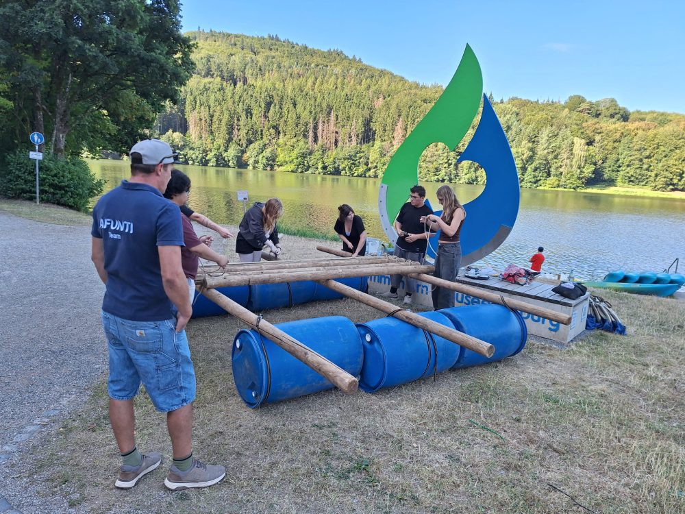 Eine Gruppe Teilnehmende bauen ein Floss aus blauen Tonnen, Holzstämmen, Seilen und Spanngurten.