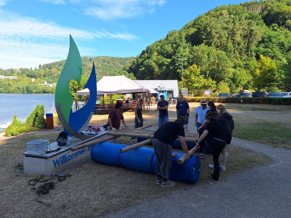 Eine Gruppe Teilnehmende bauen ein Floss aus blauen Tonnen, Holzstämmen, Seilen und Spanngurten.
