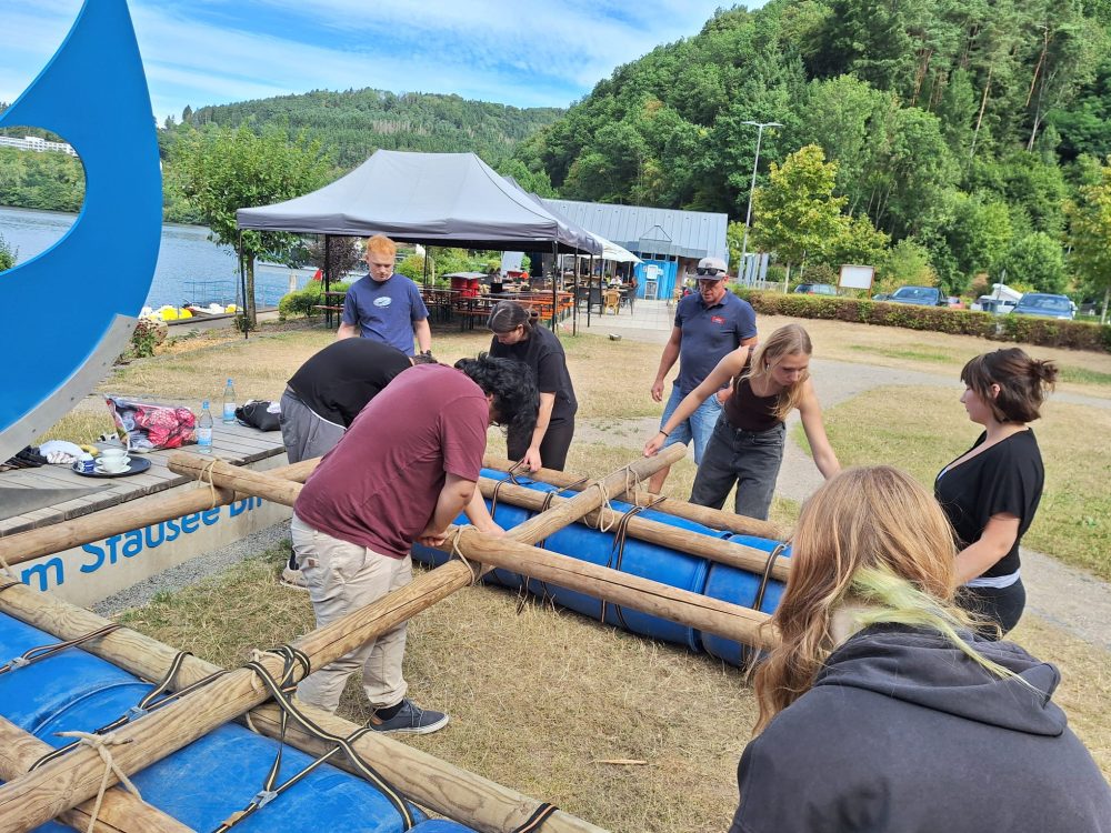 Eine Gruppe Teilnehmende bauen ein Floss aus blauen Tonnen, Holzstämmen, Seilen und Spanngurten.