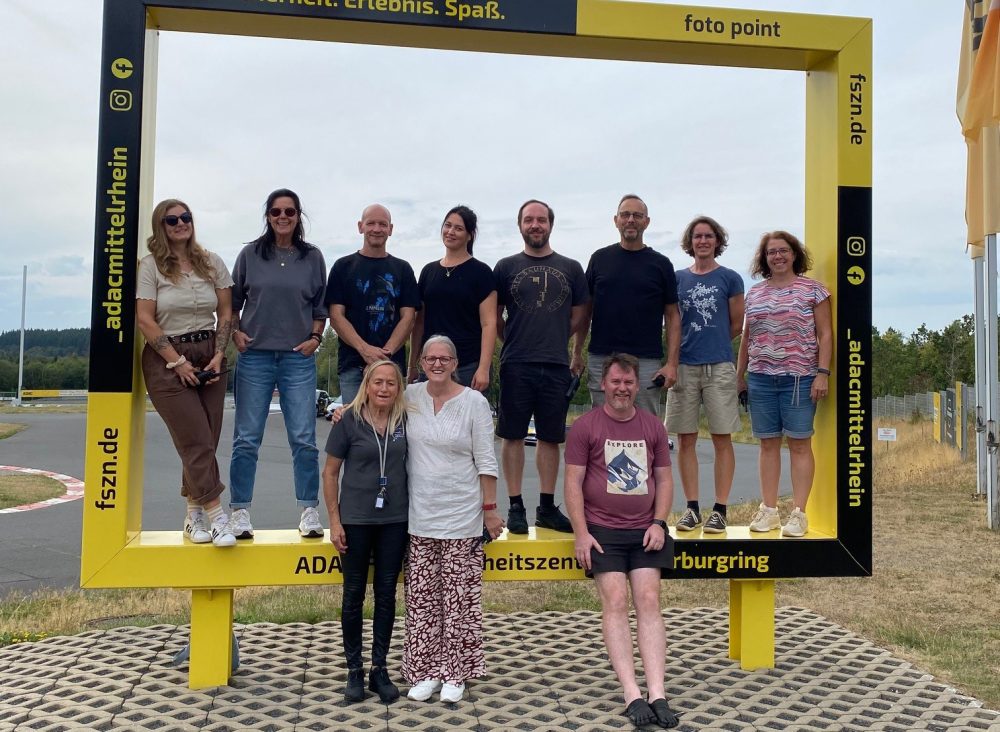 Die Gruppe der zehn Mitarbeitenden zusammen mit der Trainerin vor/in einem Foto-Rahmen "Fahrsicherheit. Erlebnis. Spaß"